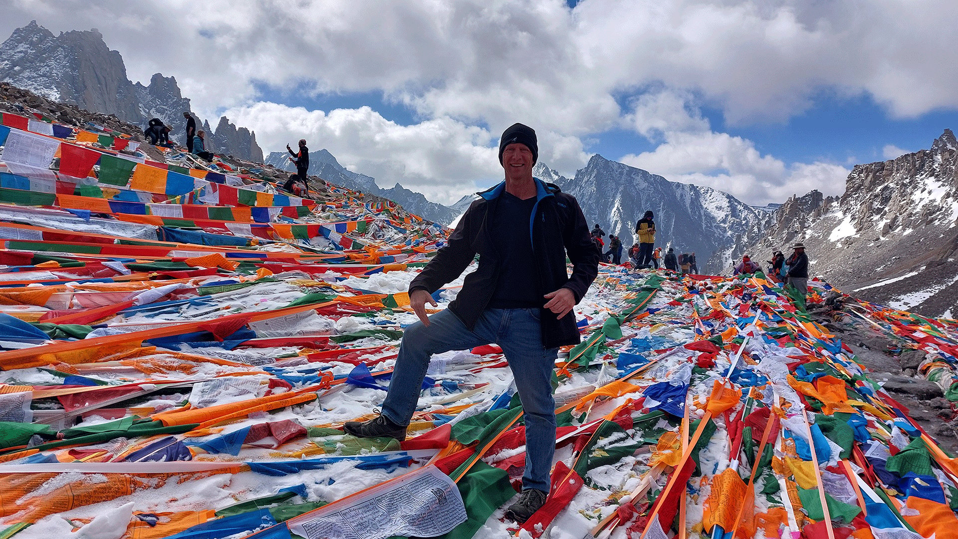 Thomas Bauer steht zwischen bunten Gebetsfahnenauf der Passhoehe Doelma La auf 5600m. Im Hintergrund sind weitere Besucherinnen und Besucher