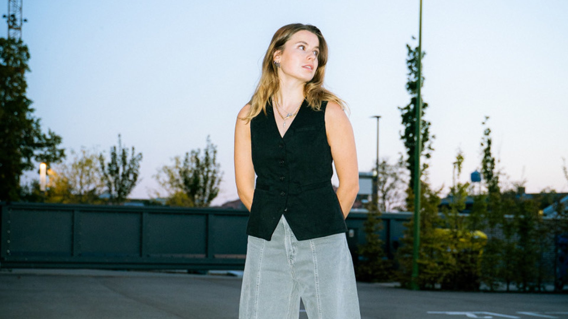 Luisa Neubauer im schwarzen ärmellosen Oberteil und heller Jeans, stehend auf einer Freifläche im Außenbereich vor Bäumen und Zaunelementen, mit seitlichem Blick vor hellem Abendhimmel