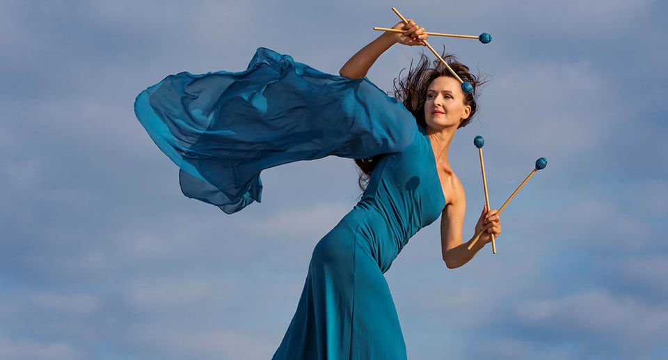 Dame in blauem Kleid mit Klöppel in Tanzbewegung vor hellblauem Hintergrund