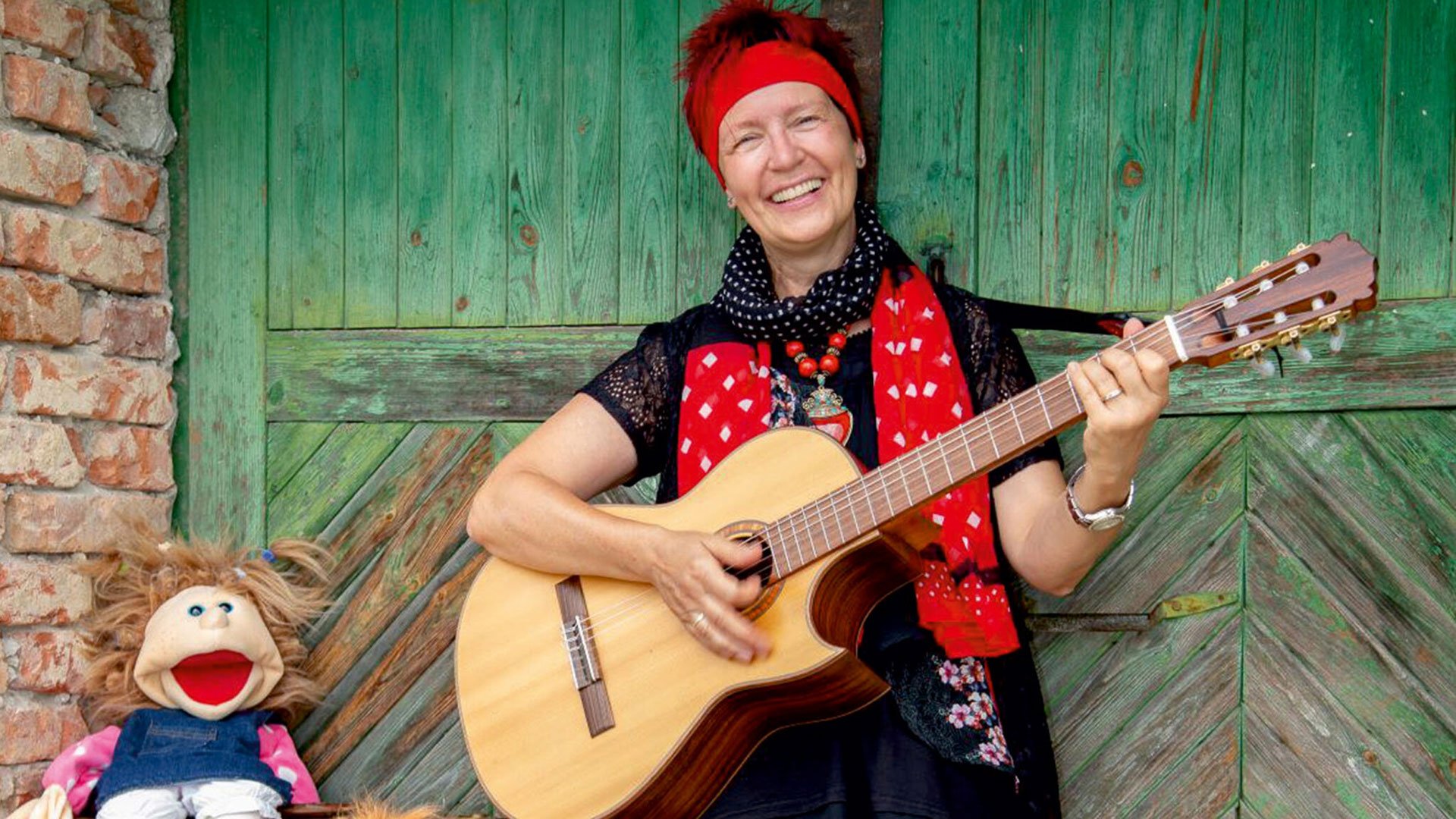 Foto zum Event von De Stianghausratschn: Die Künstlerin mit Gitarre und einer Puppe vor einer grünen Tür