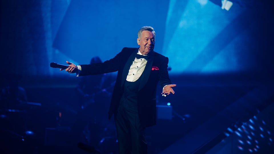 Der Künstler Roland Kaiser im schwarzen Smoking mit weißem Hemd und rotem Einstecktuch steht auf blau beleuchteter Bühne und hält ein Mikrofon dem Publikum entgegen.