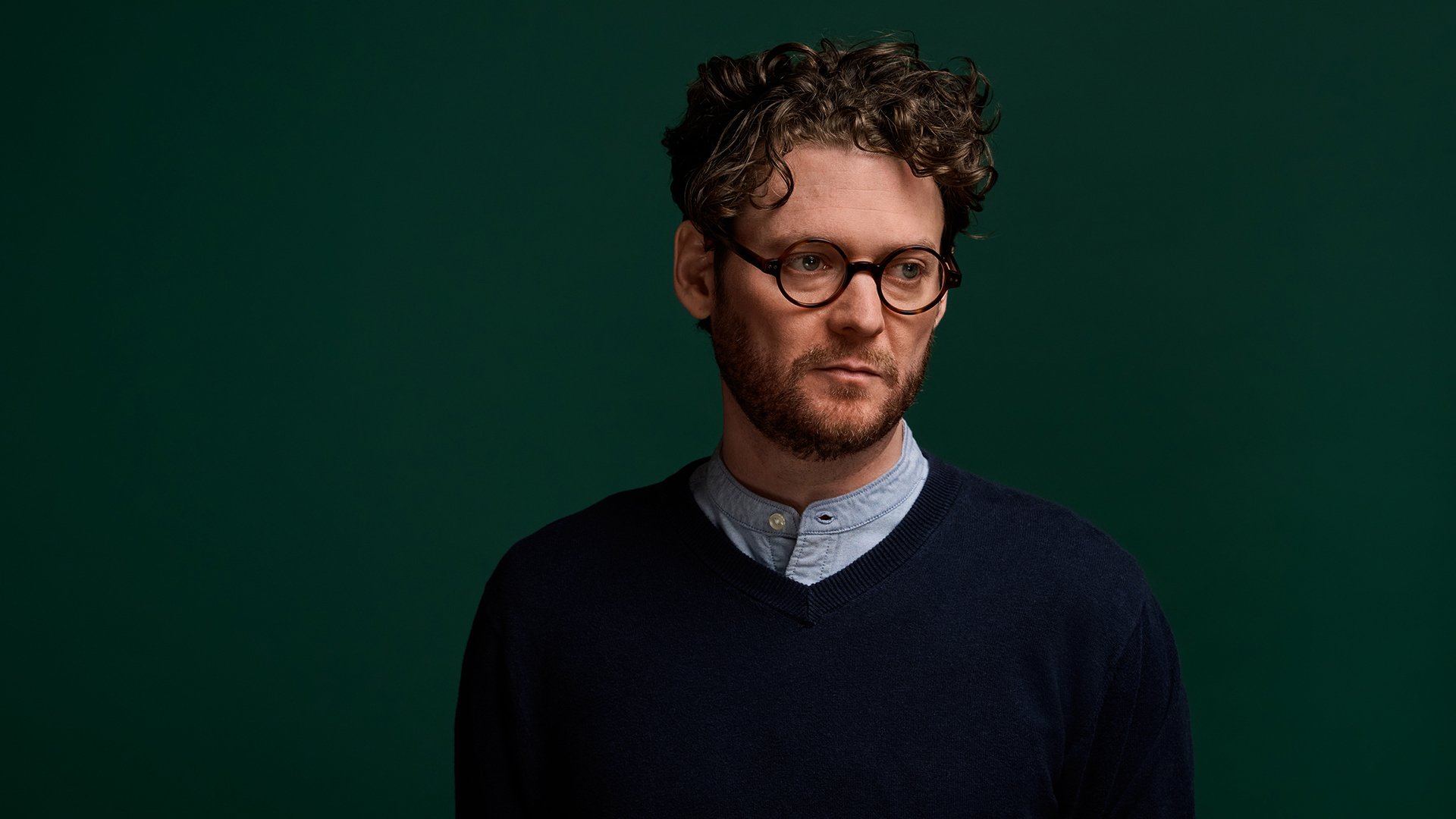 Foto von Zach Condon von der Band Beirut in dunklem Pullover undhellblauem Hemd vor dunkelgrünem Hintergrund 