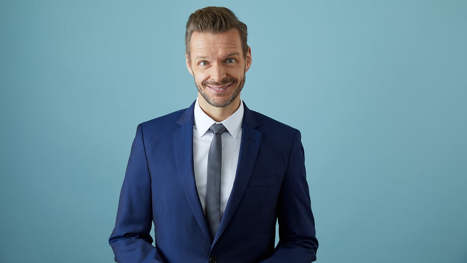 Foto für die Veranstaltung ‚Florian Schroeder – Endlich glücklich‘ mit dem Künstler im Anzug vor blauem Hintergrund.