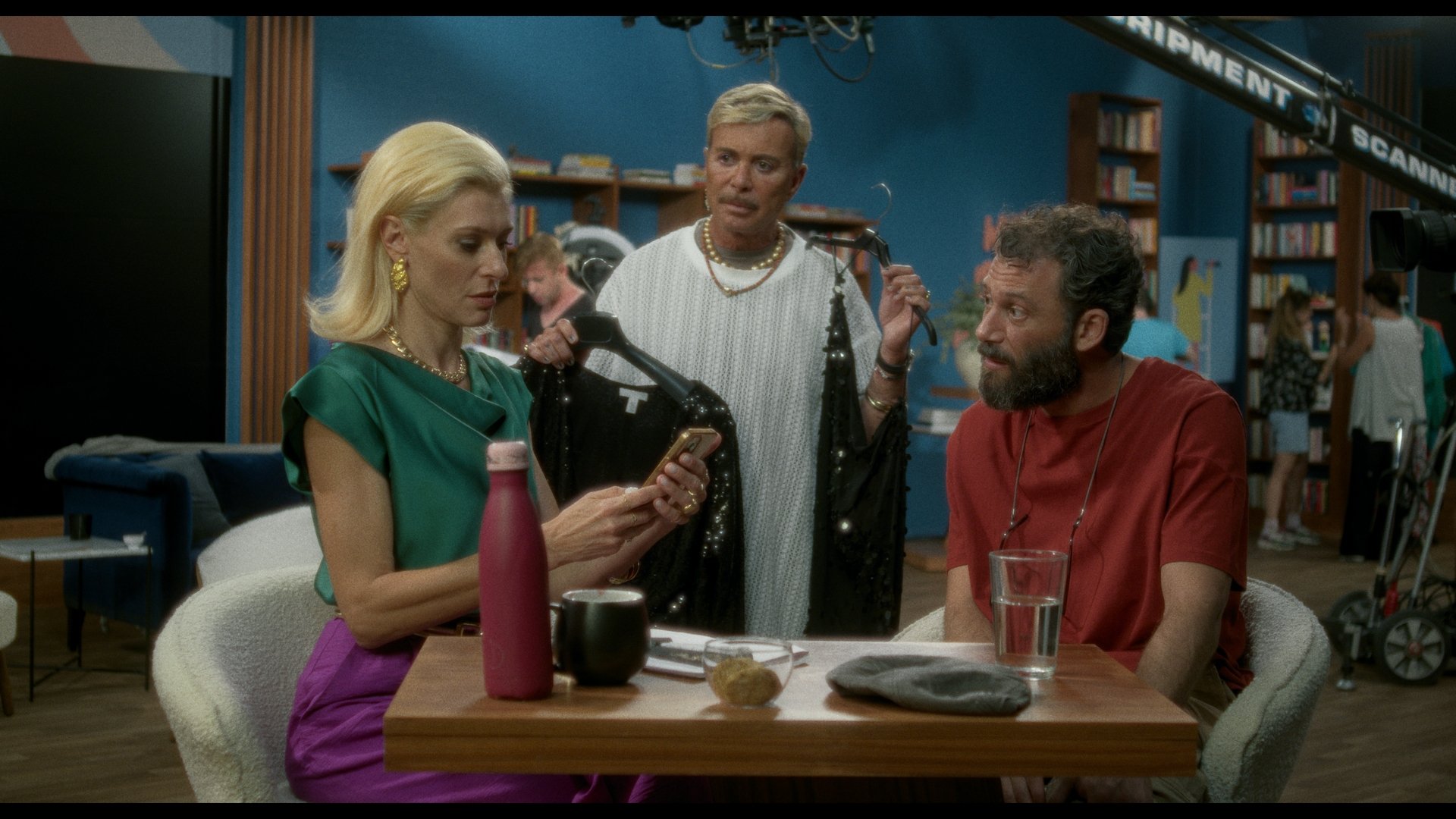 Foto vom Event ‚THE PHILOSOPHER‘ – Griechische Filmwoche München. Zwei Personen sitzen an einem Tisch, eine weitere Person steht und hält Kleidung in den Händen.