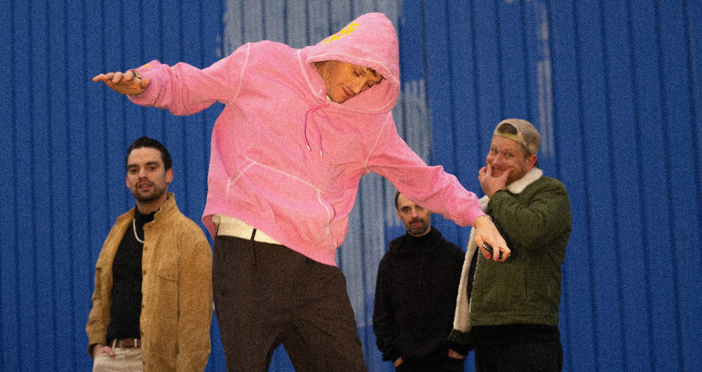 Vier Männer, einer in der Mitte mit rosa Kapuzensweatshirt springend vor blauem Hintergrund