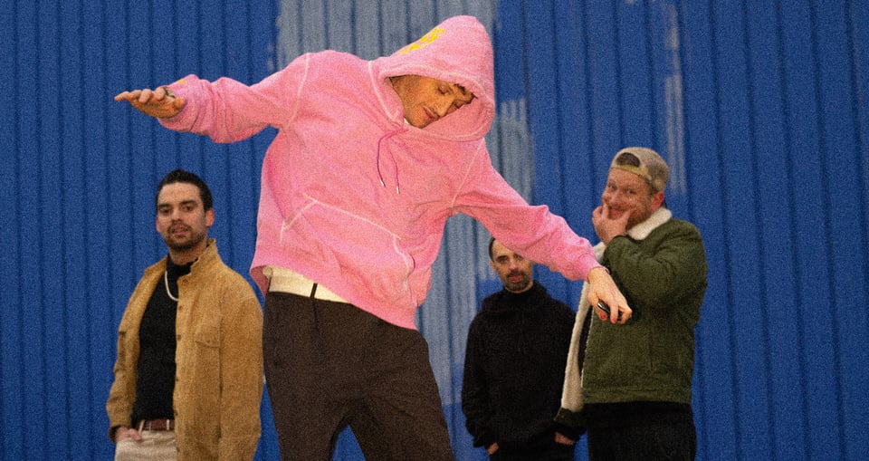 Vier Männer, einer in der Mitte mit rosa Kapuzensweatshirt springend vor blauem Hintergrund
