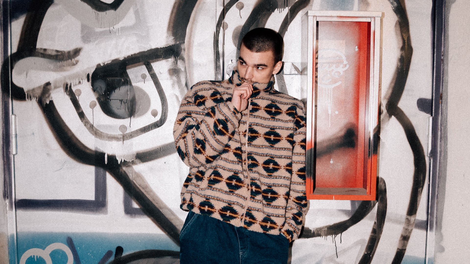 Der Künstler in einer gemusterten Jacke vor einer bunten Graffiti-Wand