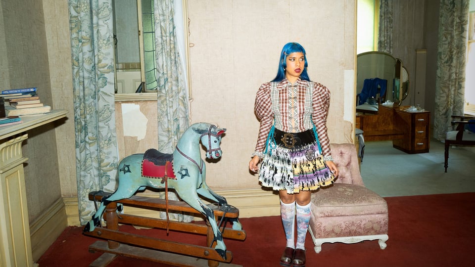 Person steht in einem vintage eingerichteten Raum neben einem hölzernen Schaukelpferd; bunte Kleidung mit gemustertem Rock und blauem Haar, im Hintergrund Möbel, Vorhänge und Spiegel.