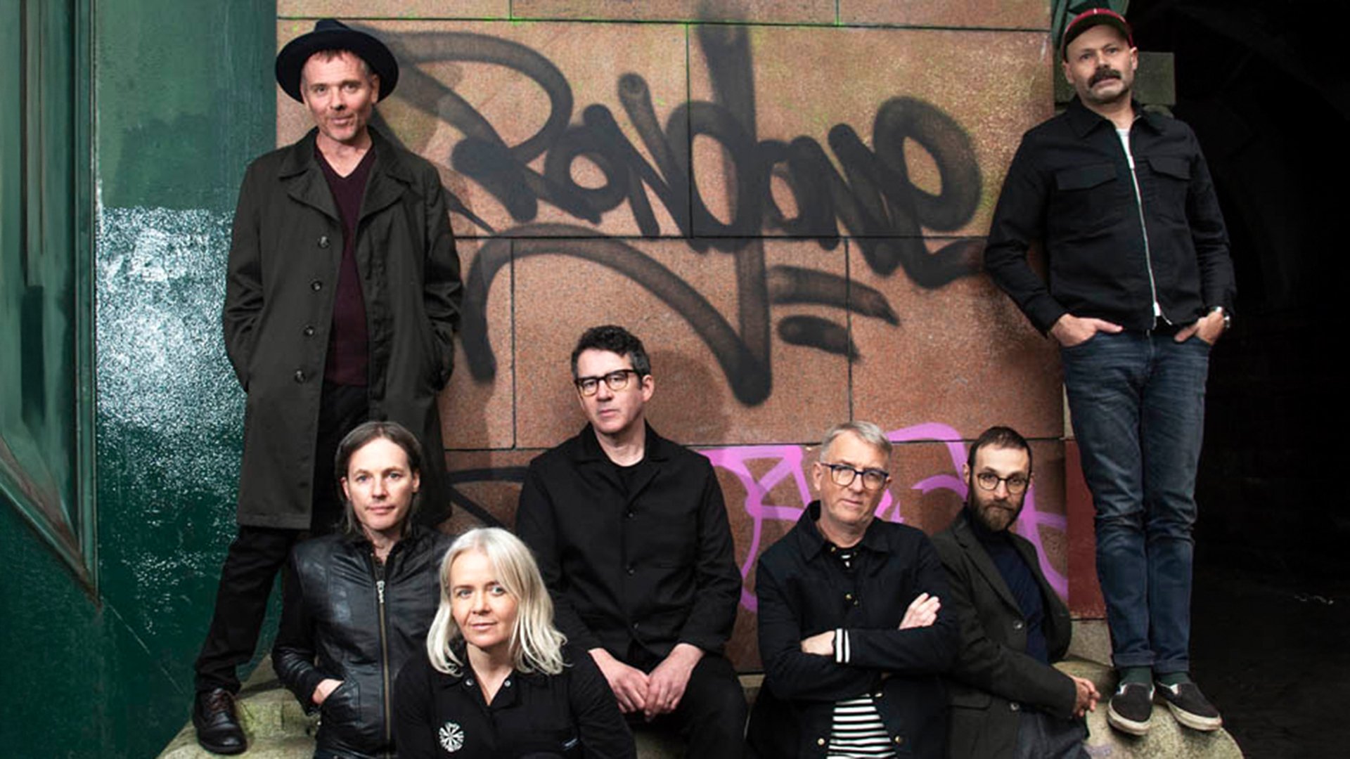 Siebenköpfige Band posiert vor einer Wand mit Graffiti; einige stehen, andere sitzen, alle tragen dunkle, legere Kleidung