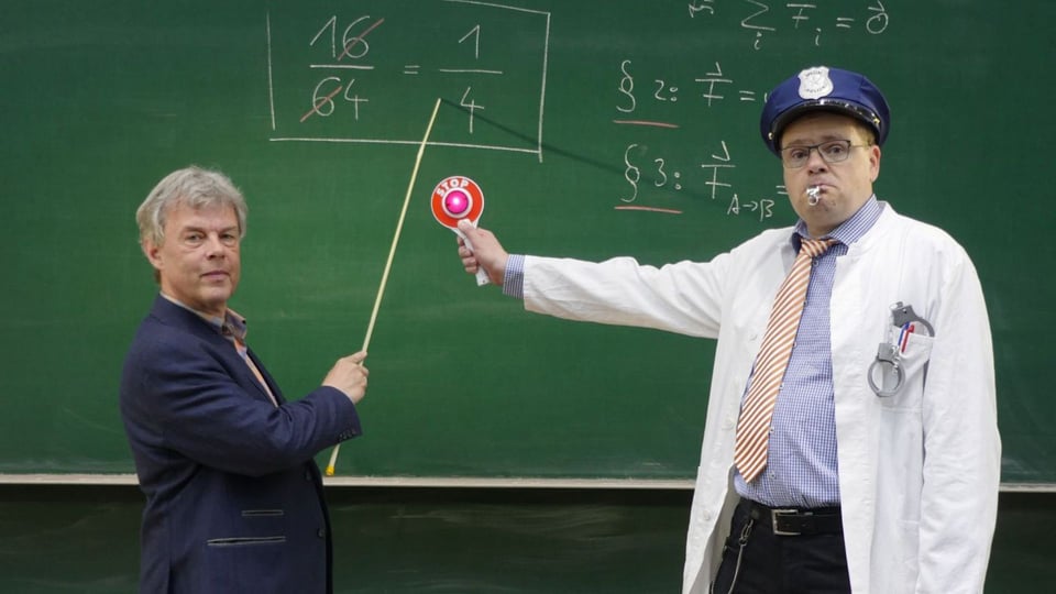 Georg Eggers und Michael Sachs stehen vor einer Tafel voller mathematischer Formeln; eine zeigt mit einem Zeigestock auf eine Gleichung, während die andere in weißem Kittel und Uniformmütze ein Halt‑Signal hochhält.
