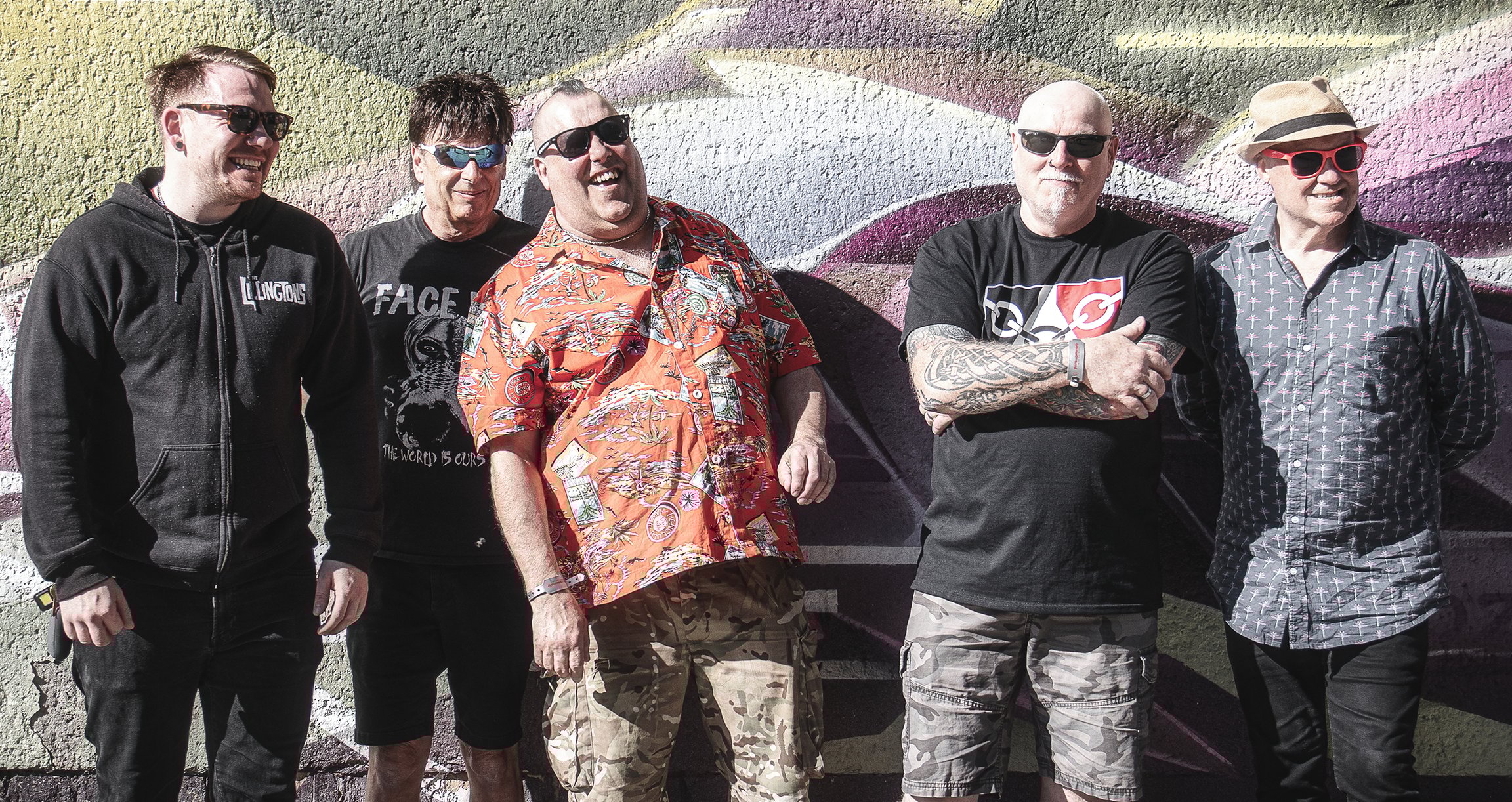 Fünf Männer stehend lachend, mit Sonnenbrille vor einer Graffitiwand.