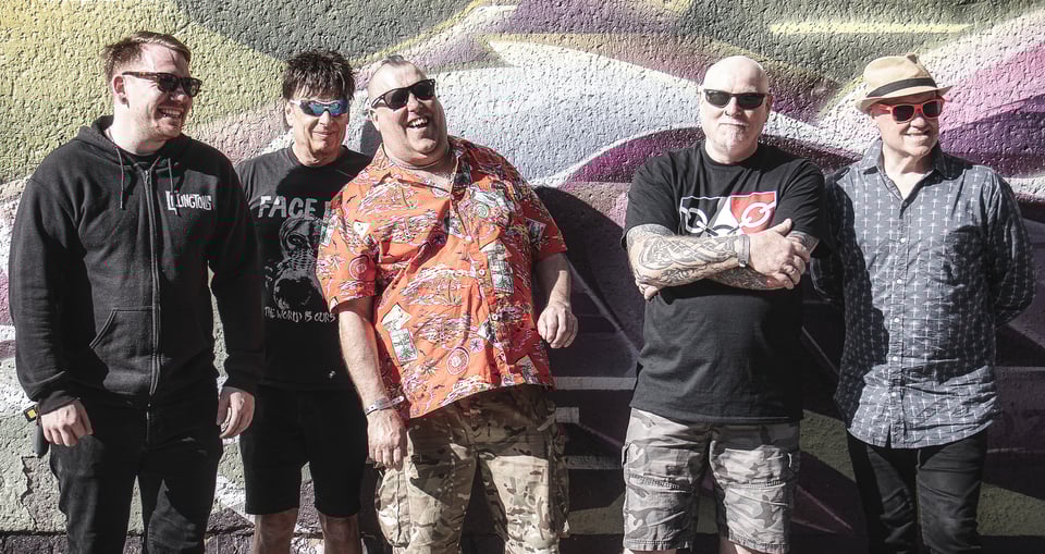Fünf Männer stehend lachend, mit Sonnenbrille vor einer Graffitiwand.