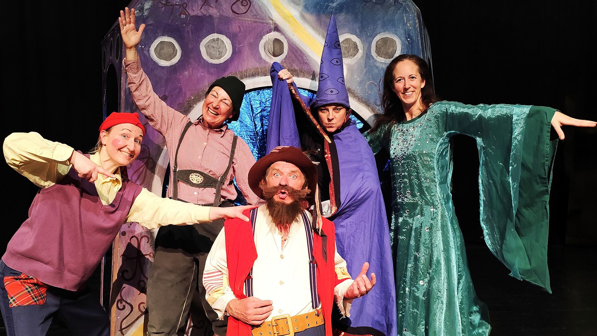 Fünf Schauspieler in bunten Kostümen spielen eine Szene aus einem Kinderstück auf einer Theaterbühne vor fantasievoller Kulisse