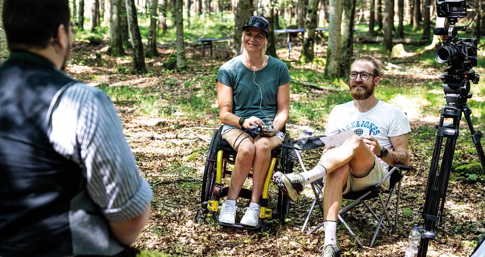 Ein Foto von drei Personen in einem Wald, wobei eine Kamera einen der Jungen filmt.