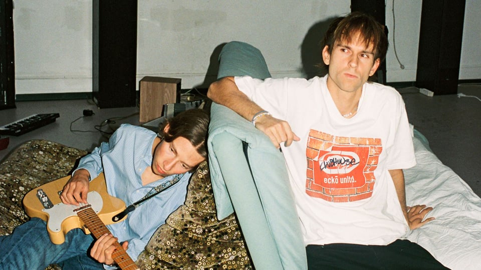 Foto für das Event Zimmer90 – Arthouse Tour 2026 mit den beiden Künstlern, die jeweils auf einem Sofa sitzen; einer von ihnen hält eine Gitarre in der Hand.