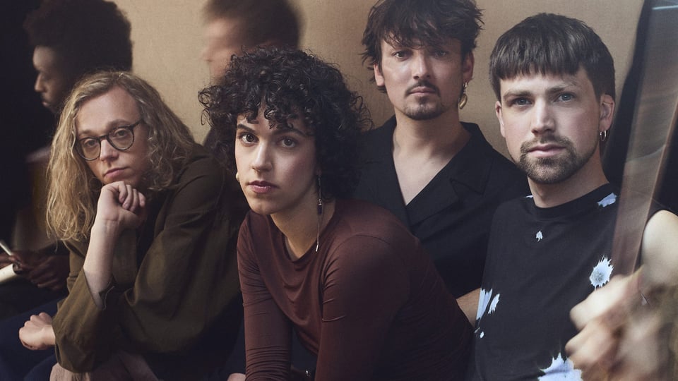 Ein Foto der Band in Freizeitkleidung nebeneinander sitzend, im Hintergrund udn seitlich verschwommene Personen