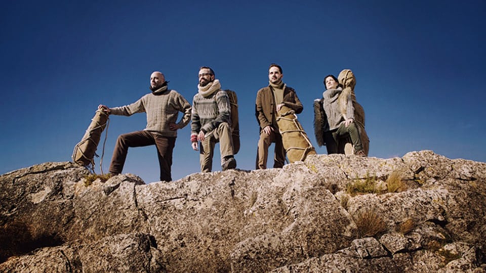 Personengruppe auf einer felsigen Anhöhe mit Instrumentenkoffern vor klarem blauem Himmel. Die Mitglieder des Quiroga Quartetts stehen in Outdoor-Kleidung auf einem Felsrücken und blicken in die Ferne