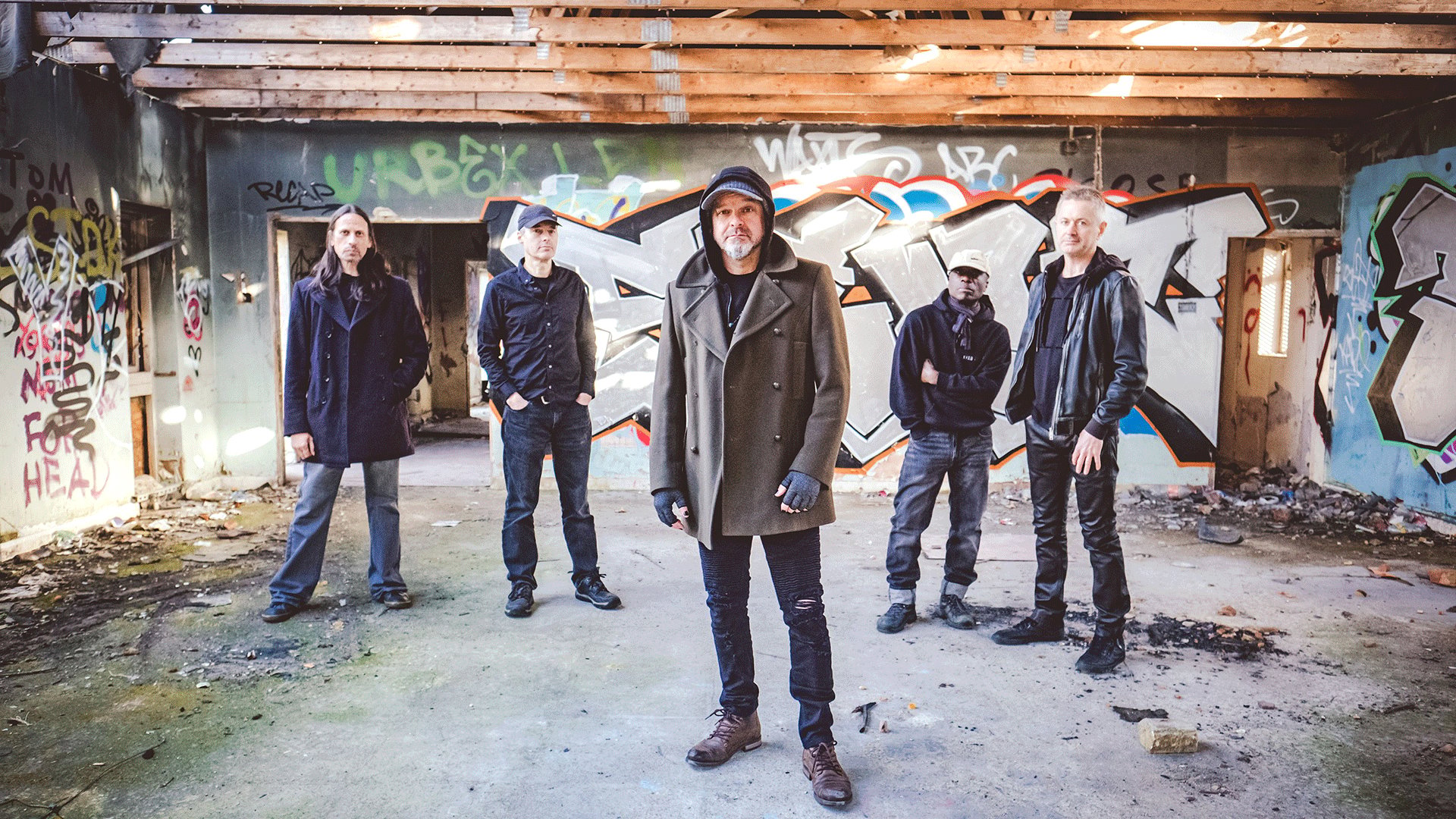Bandfoto von Threshold in einer verlassenen, mit Graffiti besprühten Industriehalle. Fünf Bandmitglieder stehen in lockerer Formation vor einer großflächigen Graffitiwand, während eine Person im Vordergrund einen dunklen Mantel mit Kapuze trägt.