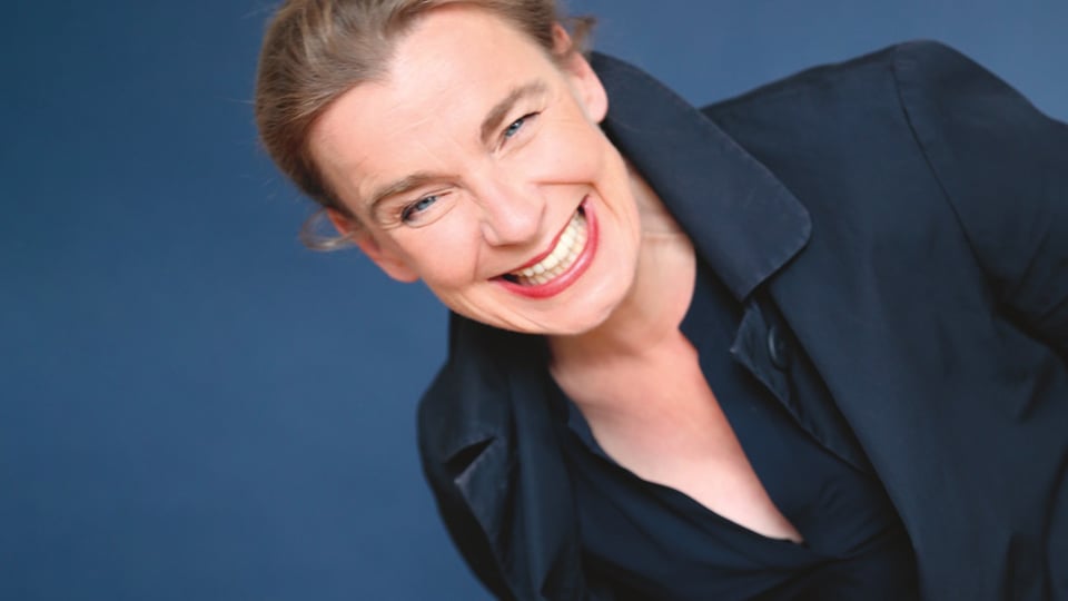 Tina Teubner lacht fröhlich vor einem blauen Hintergrund
