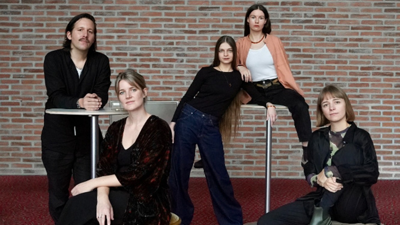 Gruppenporträt des Miriam Hanika Quartetts vor einer Backsteinwand. Fünf Personen sind in lockerer Anordnung um Stehtische positioniert und tragen überwiegend dunkle Kleidung in einer schlichten Innenraumkulisse.