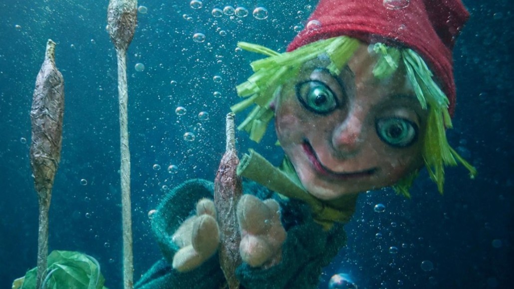 Figur des Kleinen Wassermanns mit roter Mütze und großen Augen unter Wasser zwischen Schilfpflanzen und Luftblasen. Die Puppenfigur streckt eine Hand nach vorne und ist von blaugrünem Wasser umgeben.