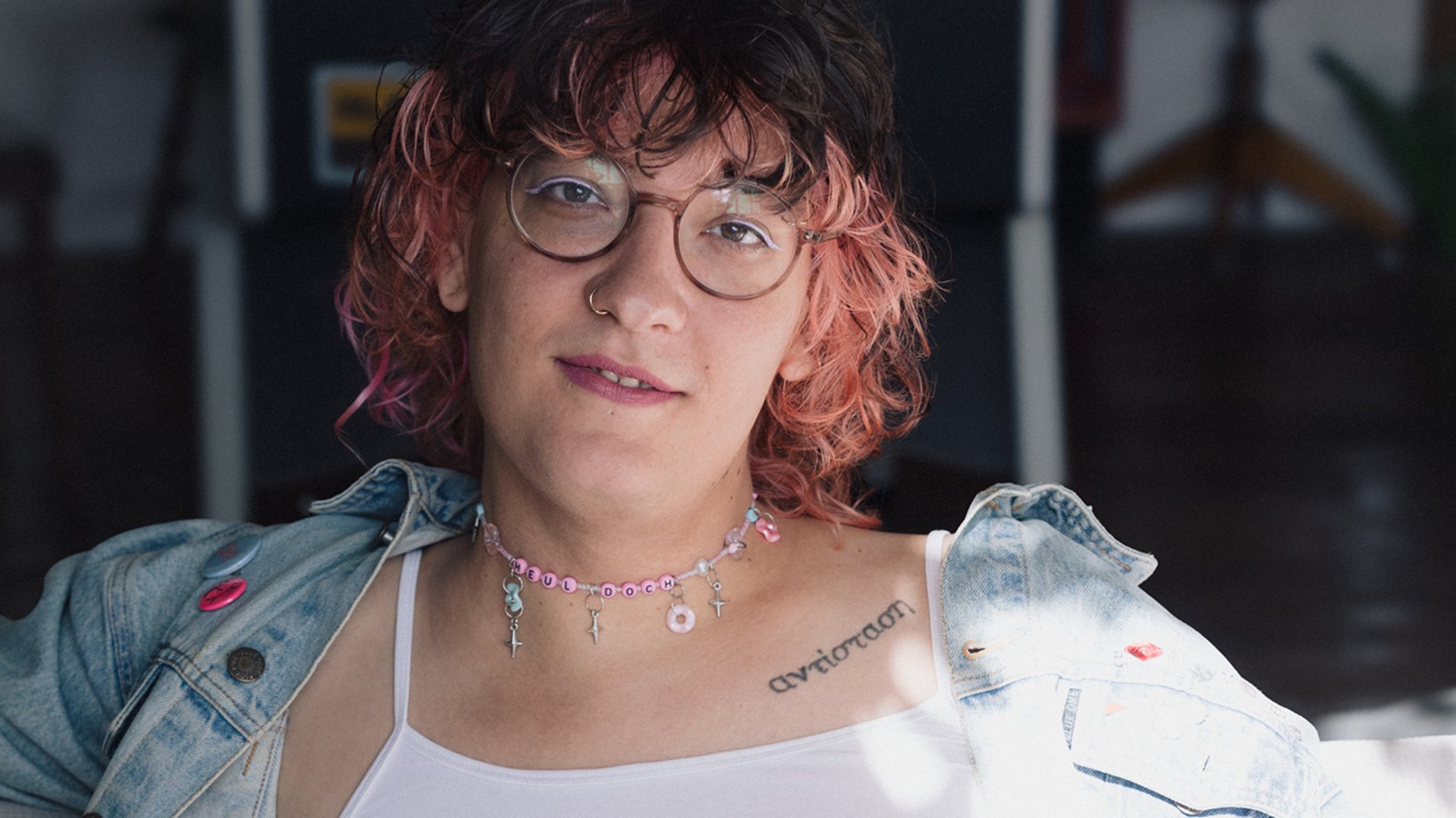 Kuku Schrapnell mit rosa schwarz gefärbtem Haar, runder Brille und heller Jeansjacke über weißem Oberteil, vor unscharfem Innenraumhintergrund mit auffälliger Halskette und sichtbarem Schriftzug-Tattoo am Schlüsselbein
