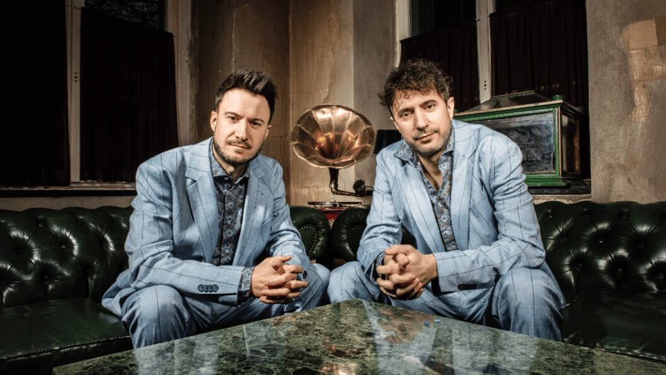 Die beiden Künstler der Ass-Dur in hellgrau gestreiften Anzügen sitzen auf dunkelgrünen Ledersofas an einem Marmortisch, im Hintergrund große Fenster und ein Grammophon