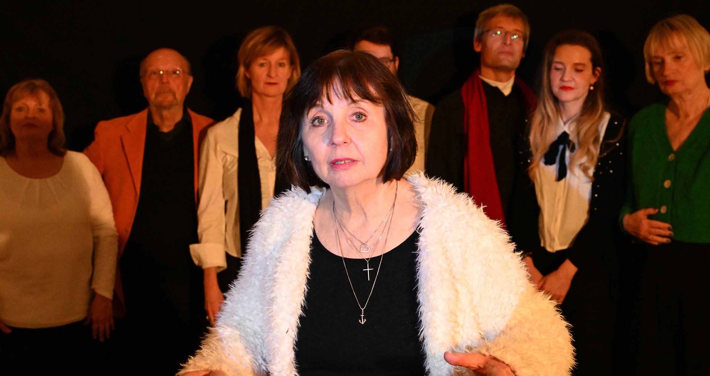 eine Dame im Vordergrund, die eine weiße Jacke trägt. Im Hintergrund eine Gruppe von Menschen mit schwarzem Hintergrund