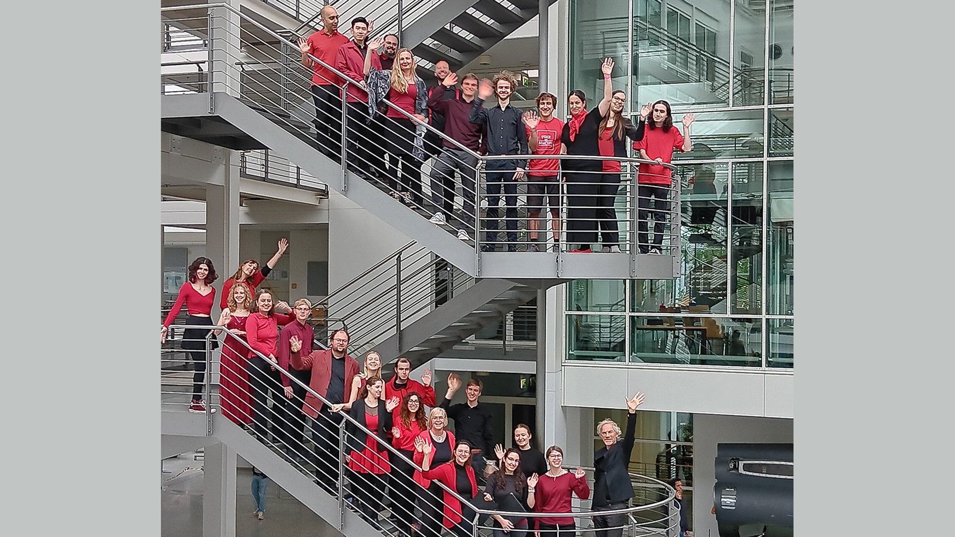 Gruppenfoto des CampusChors Garching mit zahlreichen Chormitgliedern in roten und schwarzen Outfits auf den Treppen eines modernen Gebäudes mit Glasfassade und Stahlgeländern, viele winken von den verschiedenen Ebenen des offenen Treppenhauses in die Kamera.