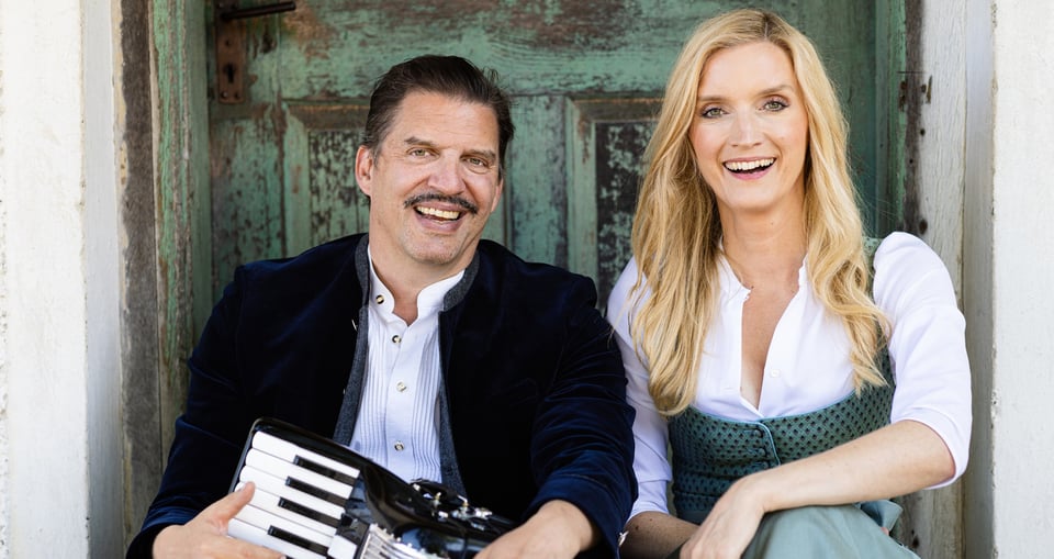 Foto einer Frau und eines Mannes mit einem Instrument in der Hand – beide in traditioneller bayerischer Tracht.