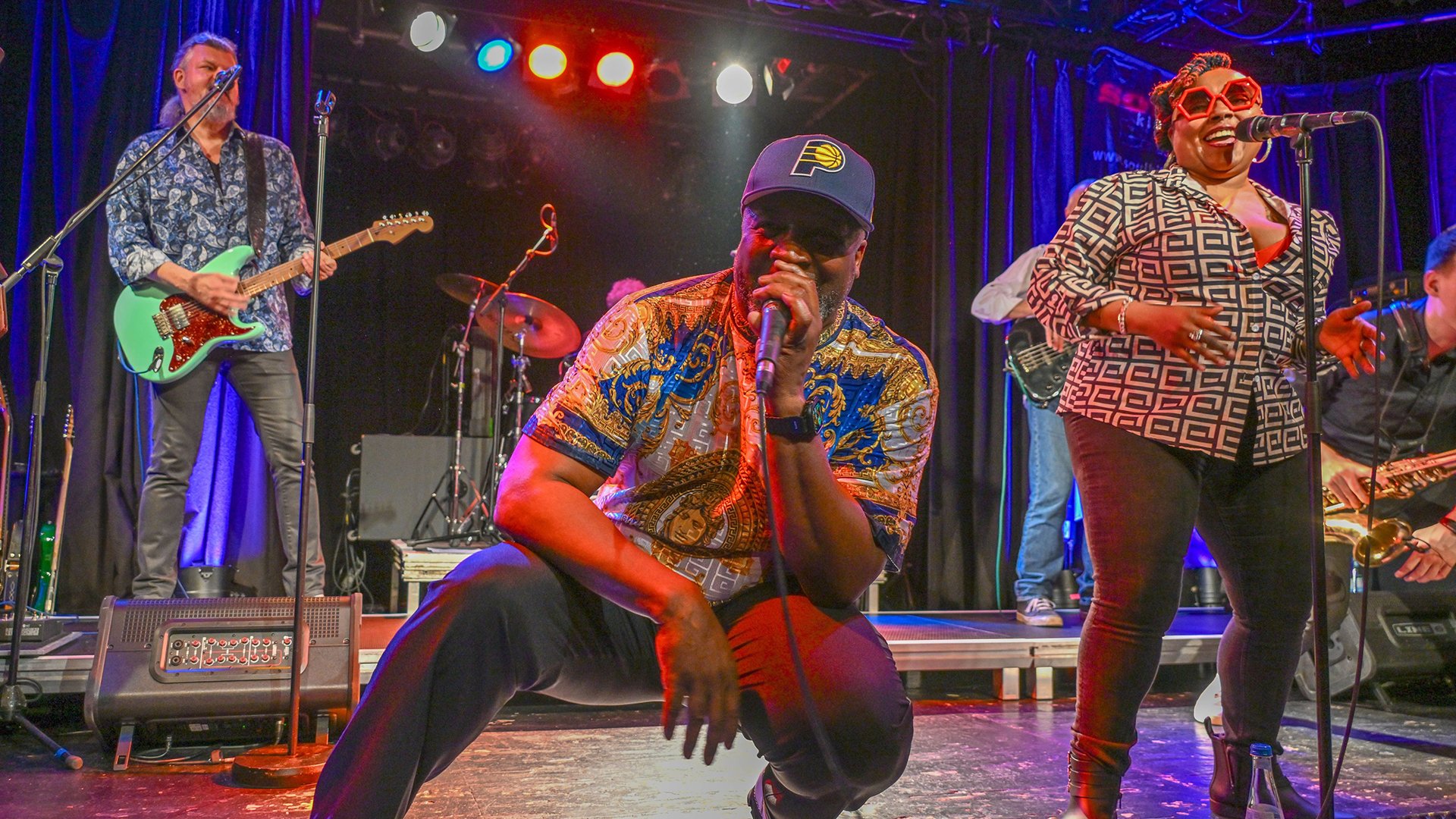 Die Band Soul Kitchen in buntem Licht und bunter Kleidung auf der Bühne mit Instrumenten, der Sänger kniet vorne im Bild