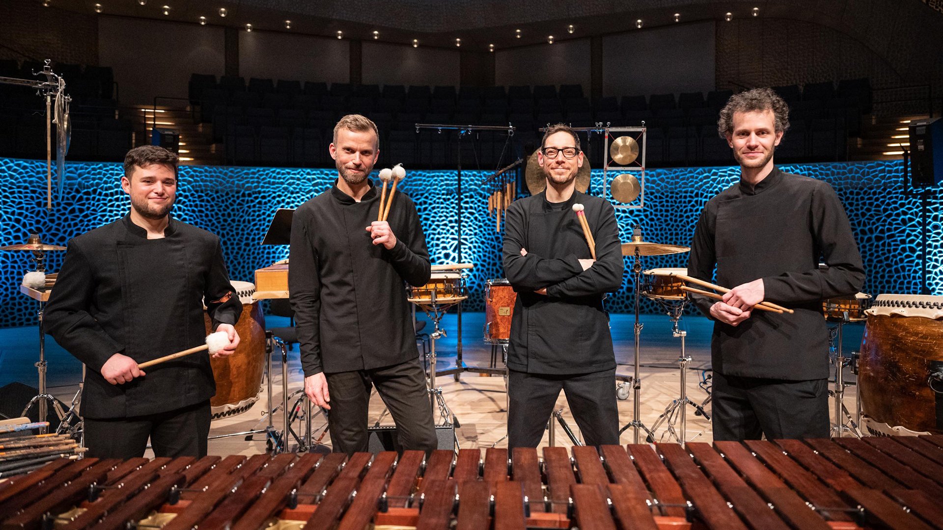 Vier Percussionisten stehen zwischen verschiedenen Schlaginstrumenten auf einer Konzertbühne und halten Schlägel in den Händen