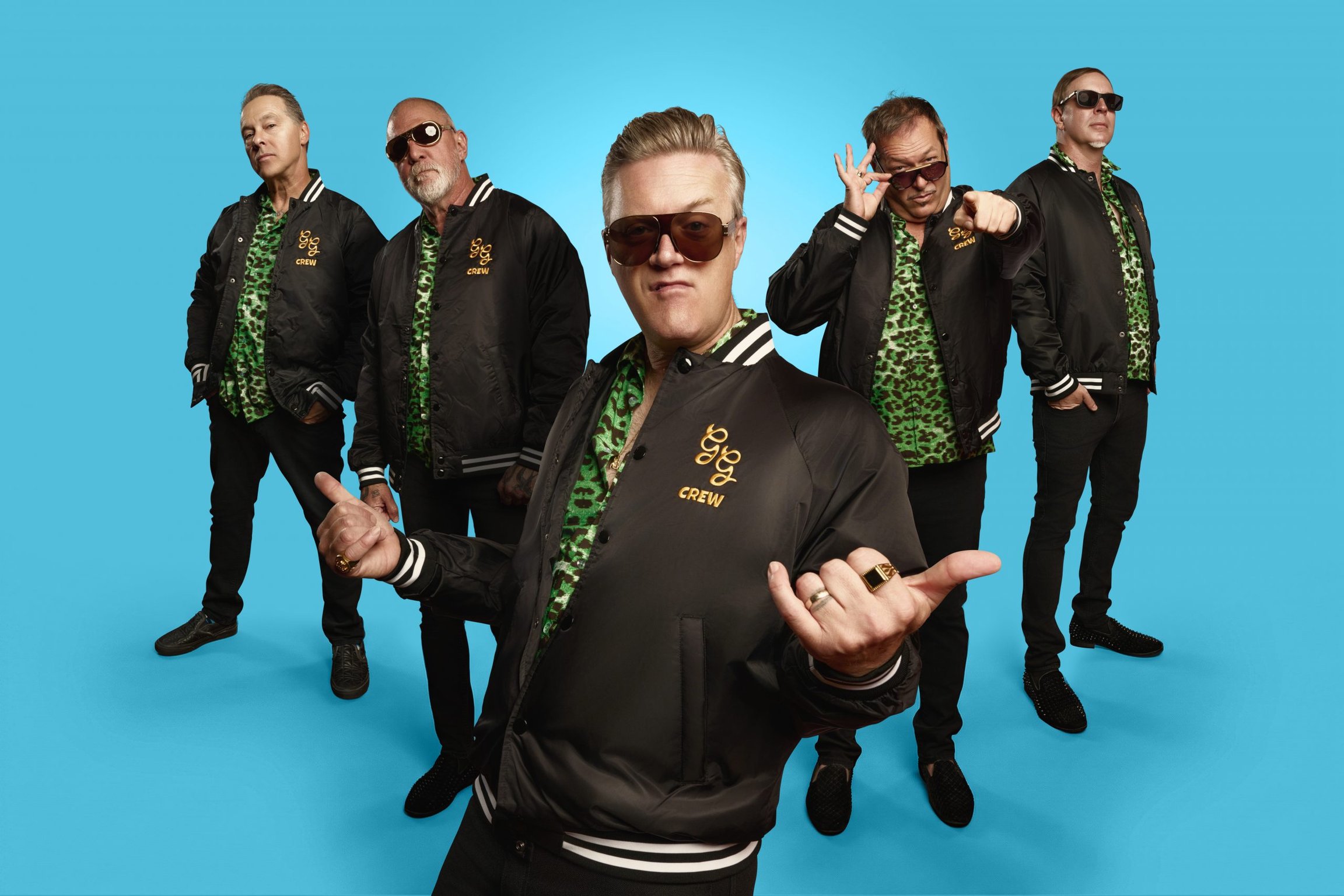 Studiofoto von fünf Bandmitgliedern der Spike and the Gimme Gimmes in schwarzen Jacken und grün gemusterten Hemden vor blauem Hintergrund, eine Person im Vordergrund mit Sonnenbrille und Handgeste
