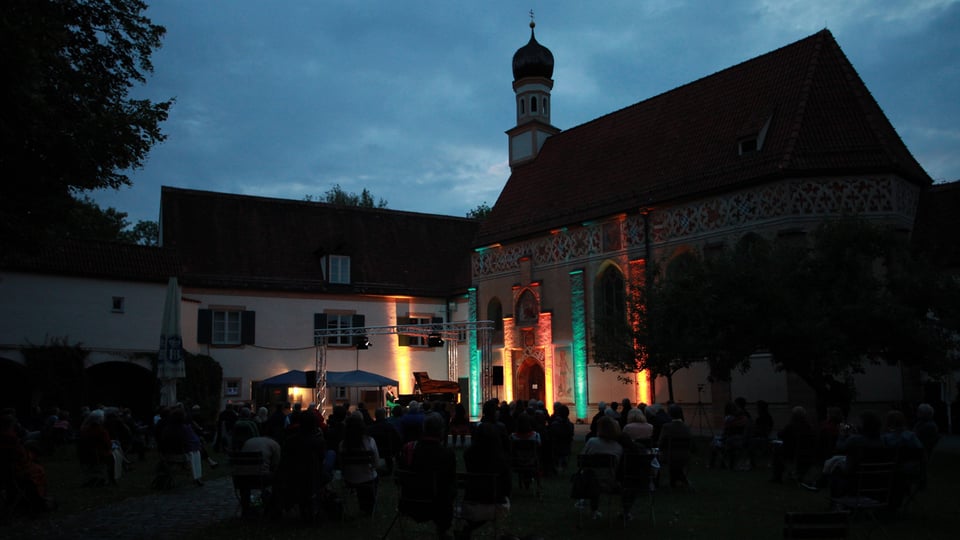 Abendliche Open‑Air‑Veranstaltung vor einem historischen Gebäude mit Kirchturm: Im Vordergrund sitzen zahlreiche Besucherinnen und Besucher auf Stühlen vor einer beleuchteten Bühne. Die Fassade des Gebäudes ist mit farbiger Lichtinszenierung in Grün‑, Orange‑ und Warmtönen angestrahlt. Auf der Bühne sind Lichttechnik und ein Klavier erkennbar. Der Himmel zeigt dunkle Abendwolken, Bäume rahmen die Szene ein. Das Bild vermittelt eine stimmungsvolle Konzert‑ oder Kulturveranstaltung im Außenbereich.