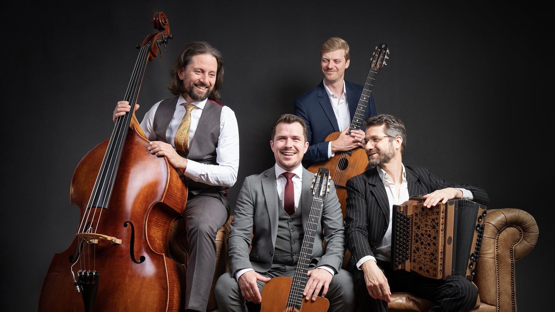 Vier Herren in klassischen Anzügen sitzen mit Instrumenten lachend auf einem braunen Ledersofa vor dunklem Hintergrund