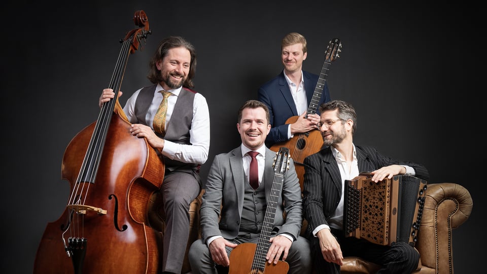 Vier Herren in klassischen Anzügen sitzen mit Instrumenten lachend auf einem braunen Ledersofa vor dunklem Hintergrund