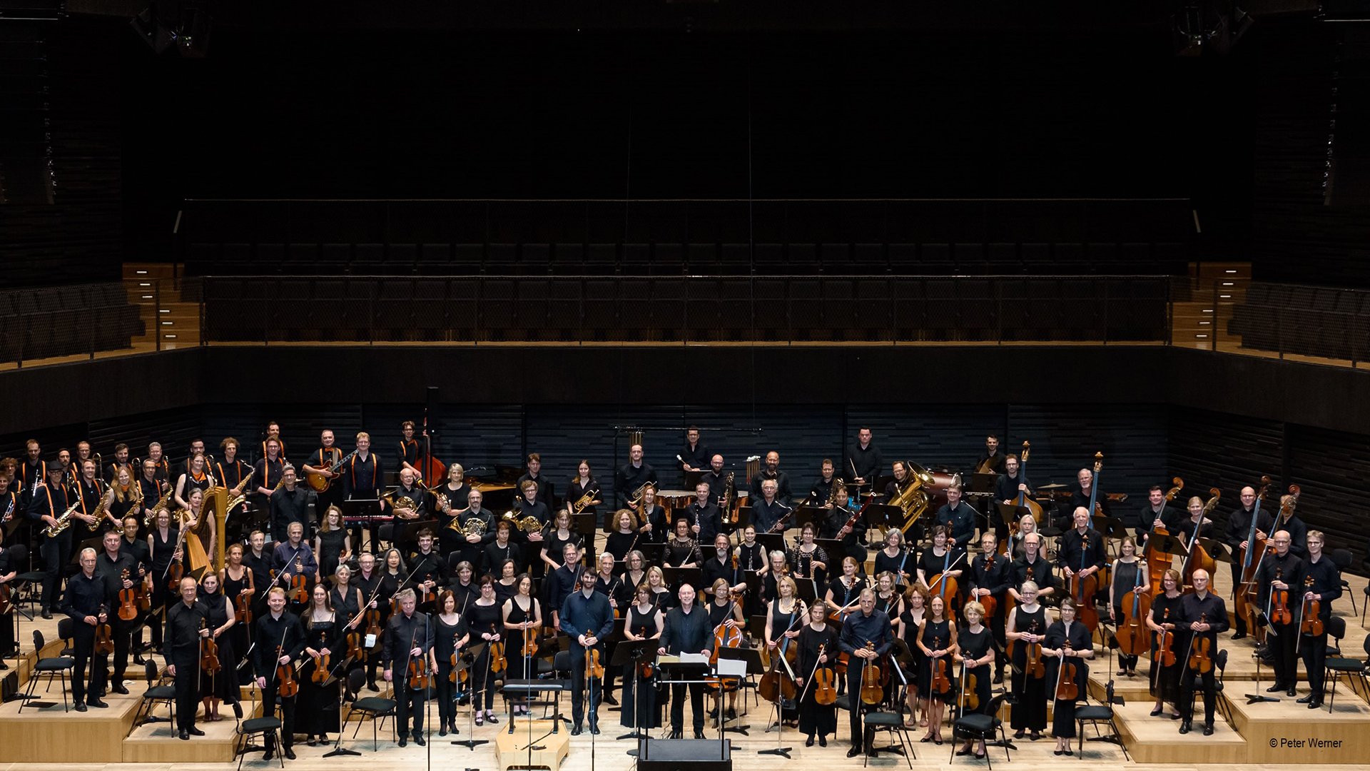 Großes Orchester mit Musiker:innen und Instrumentalist:innen auf der Bühne eines modernen Konzertsaals. Die Ensemblemitglieder stehen in schwarzer Konzertkleidung zwischen Streich-, Blas- und Schlaginstrumenten.