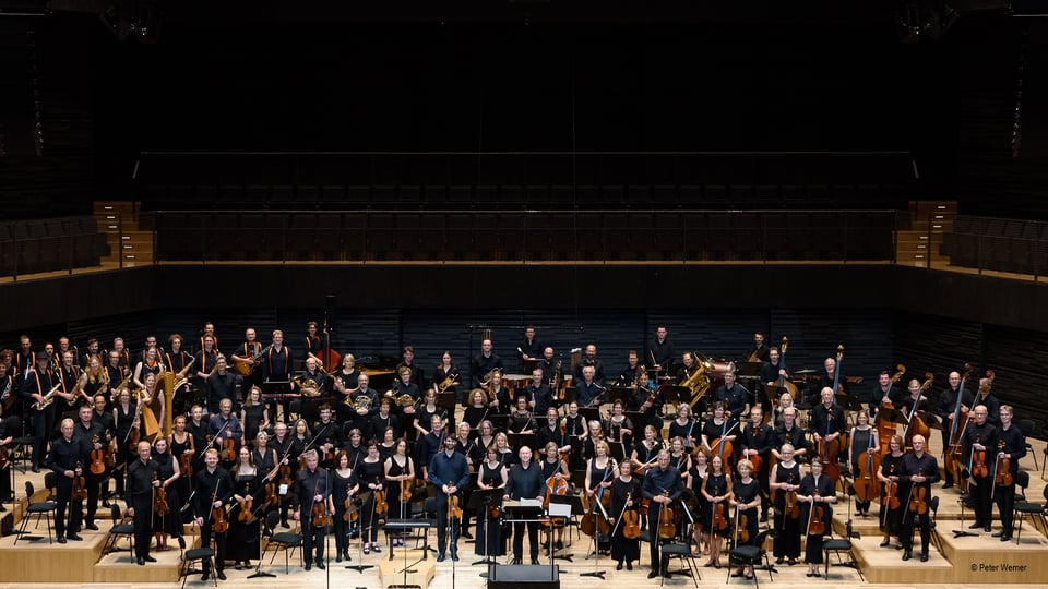 Großes Orchester mit Musiker:innen und Instrumentalist:innen auf der Bühne eines modernen Konzertsaals. Die Ensemblemitglieder stehen in schwarzer Konzertkleidung zwischen Streich-, Blas- und Schlaginstrumenten.
