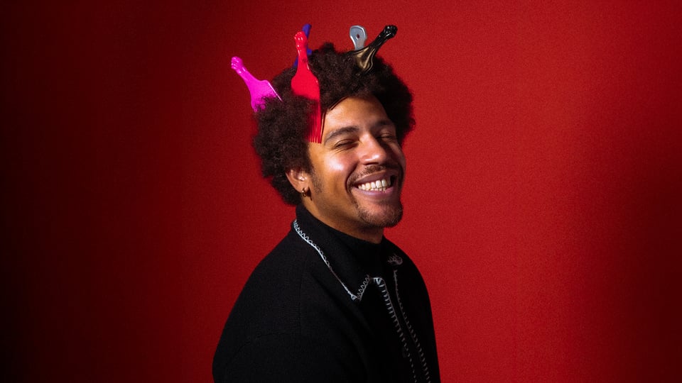 Foto für die Veranstaltung NoMBe mit dem Künstler, dessen Afro-Haarfrisur mit einigen eingesteckten Kämmen zu sehen ist, vor rotem Hintergrund.