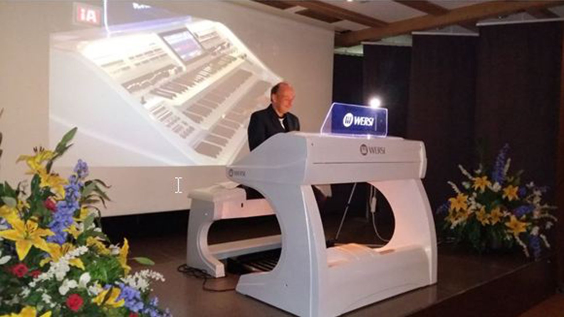 Heiko Schneider spielt an einer modernen weißen Orgel auf einer Bühne, im Hintergrund eine projizierte Orgelansicht und Blumenarrangements