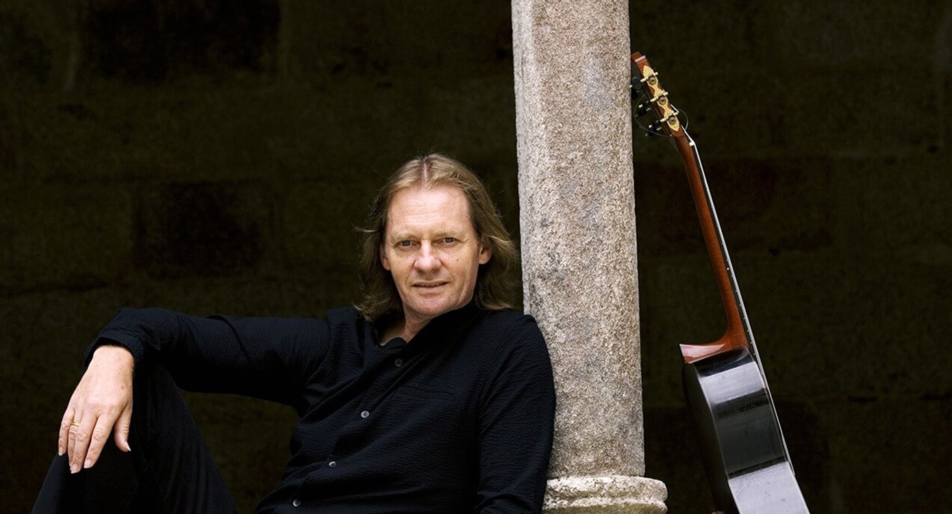 David Russell stützt sich auf eine Säule und eine Gitarre vor einem schwarzen Hintergrund