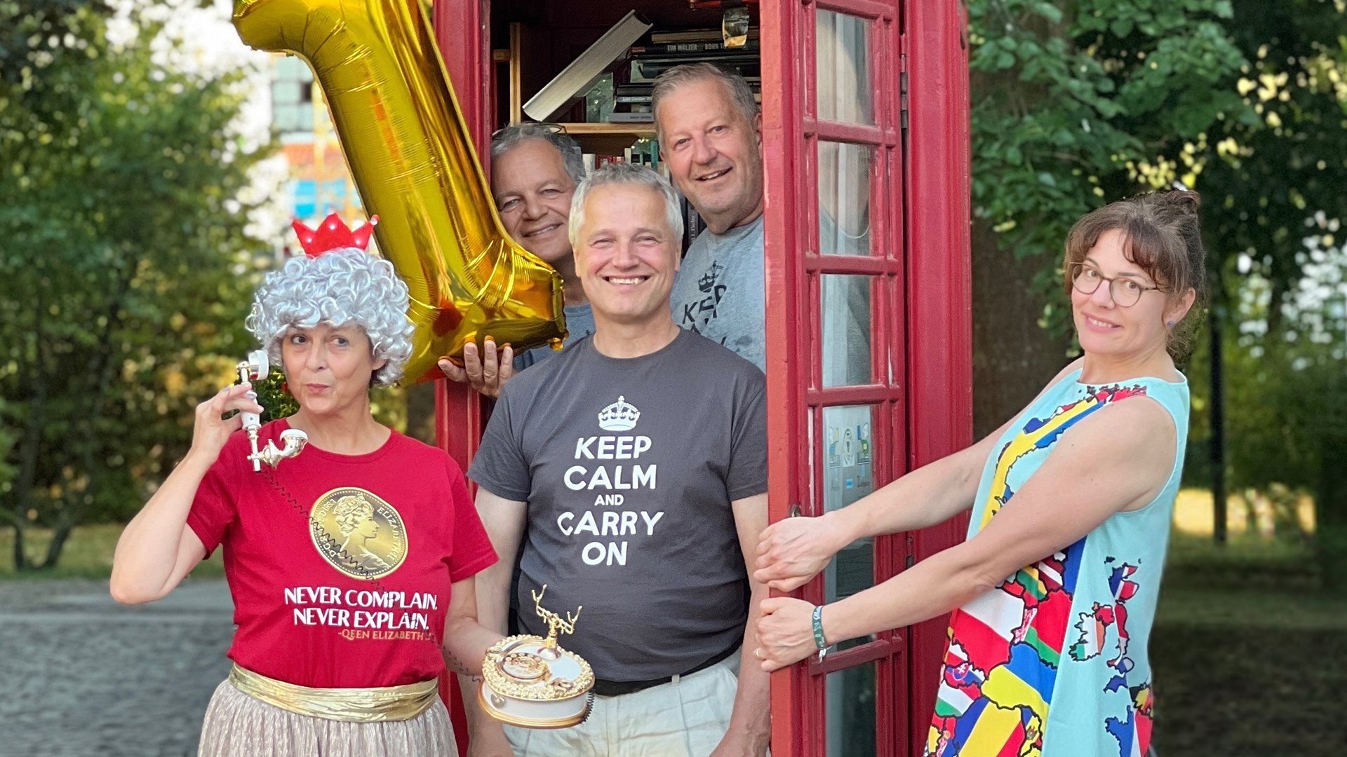 Gruppe von fünf Personen vor einer roten Telefonzelle im Freien mit Ballons in einer Parkumgebung