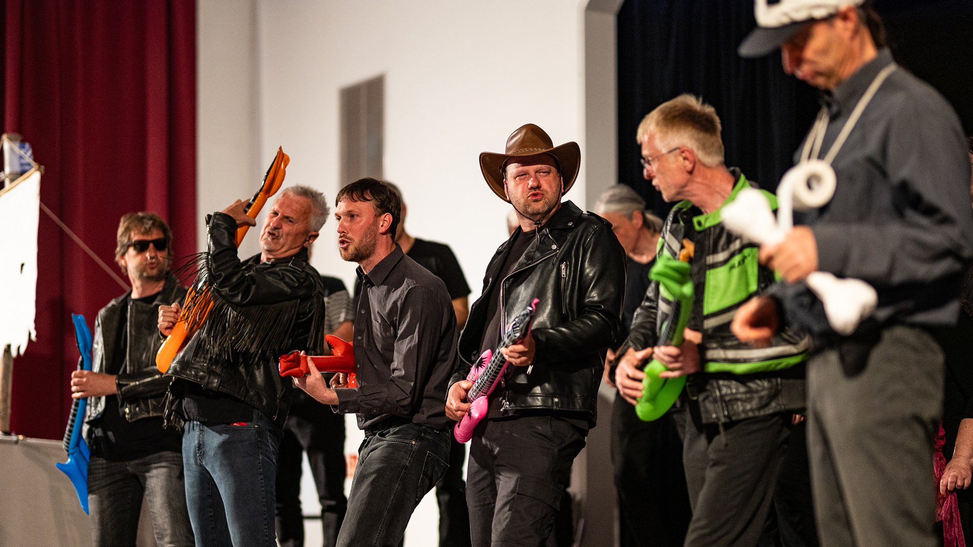 Mehrere Personen stehen auf einer Bühne und spielen mit aufblasbaren Spielzeuginstrumenten. Einige tragen Kostüme, darunter eine Cowboyhut- und Lederjackenkombination