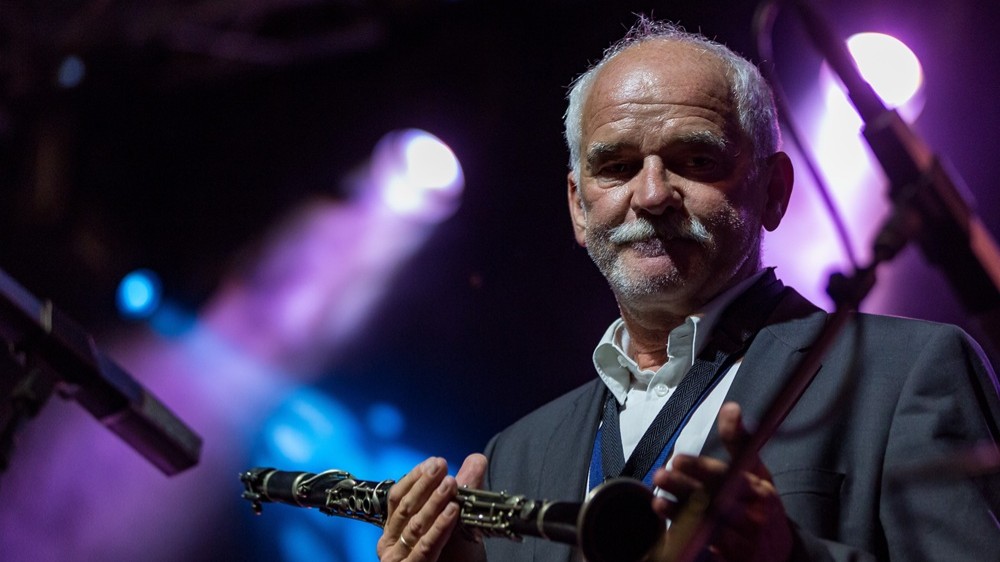 Klarinettist steht auf einer Bühne mit Bühnenbeleuchtung und hält eine Klarinette in den Händen, Nahaufnahme eines Musikers im Anzug vor unscharfem Hintergrund mit Scheinwerfern.