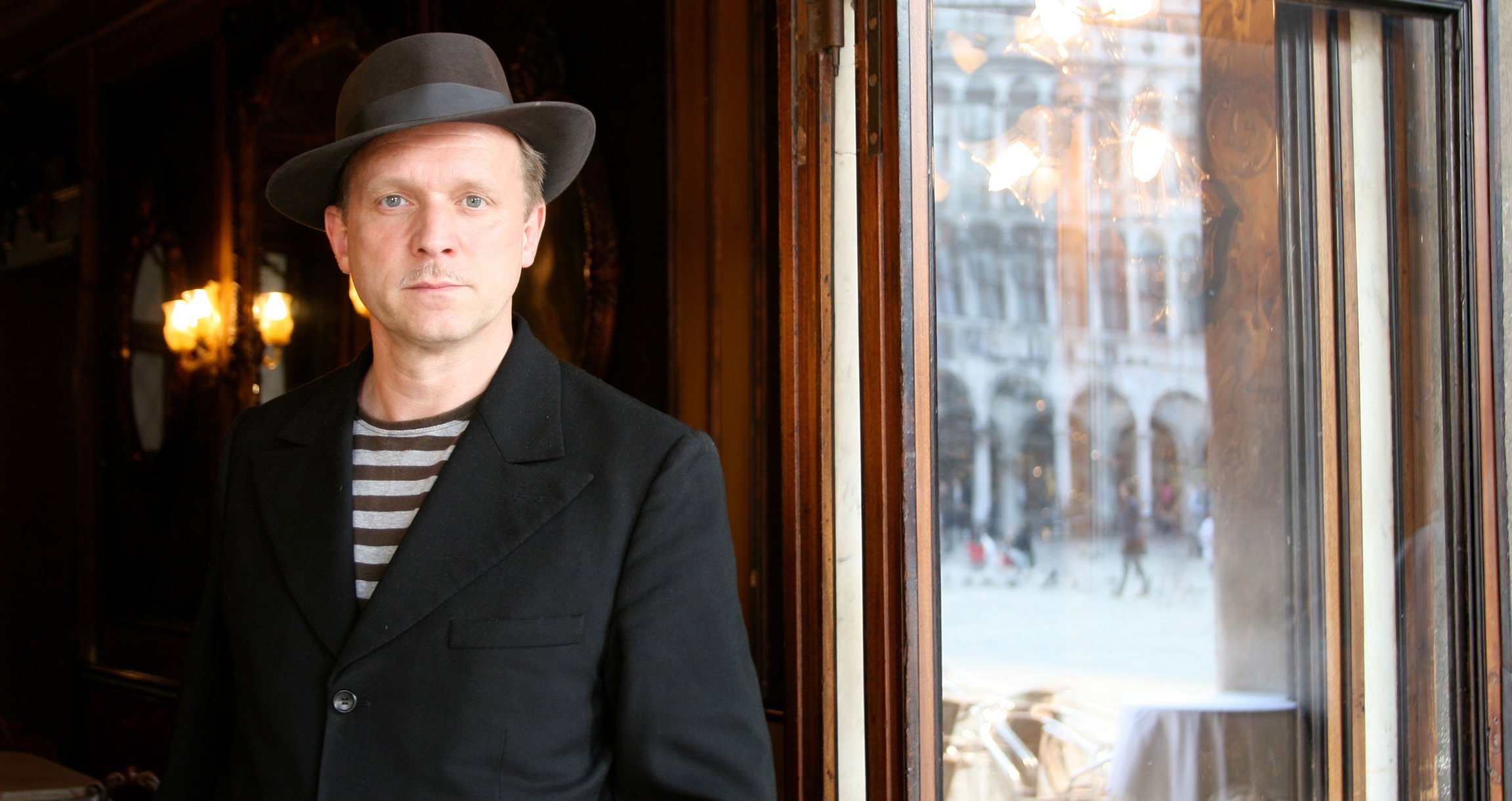 Mann mit gestreiften Shirt, schwarzem Sakko und schwarzem Hut vor antiker Fensterfassade