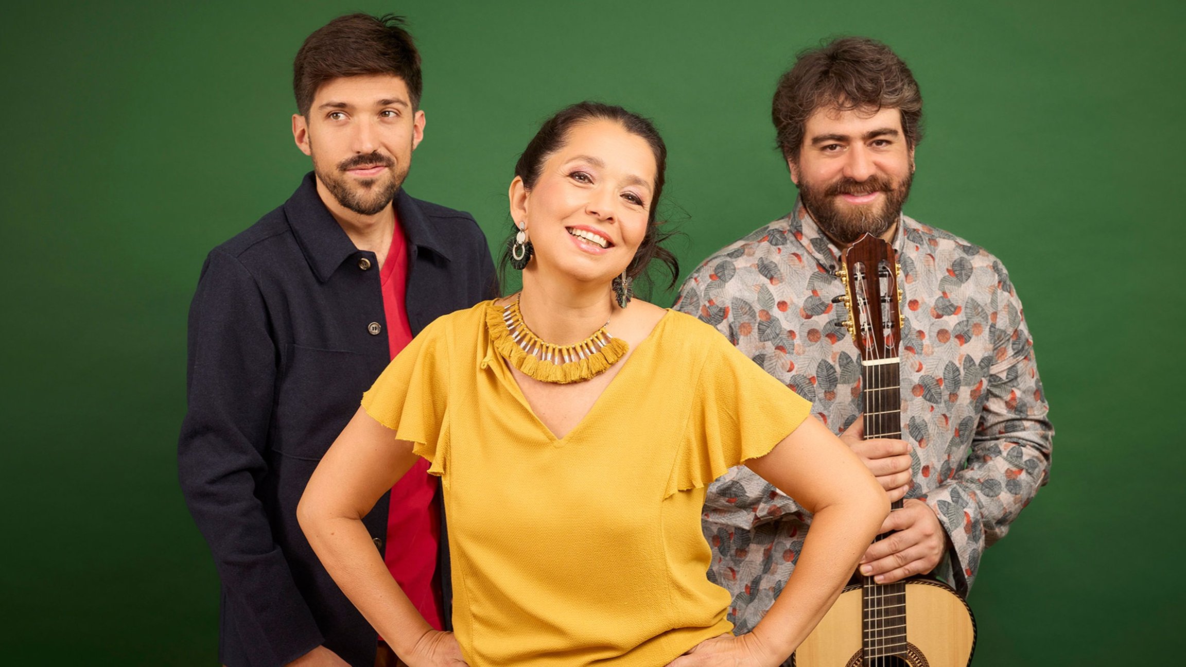 Studiofoto mit drei Personen der Gruppe Amaré vor grünem Hintergrund, eine Person vorne in gelbem Oberteil, daneben zwei weitere Personen, eine mit Gitarre in der Hand