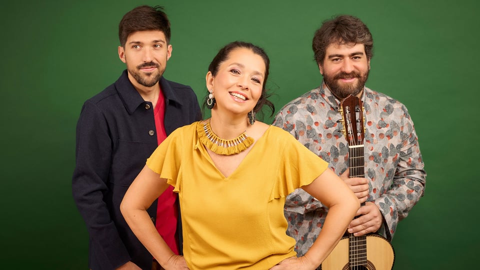 Studiofoto mit drei Personen der Gruppe Amaré vor grünem Hintergrund, eine Person vorne in gelbem Oberteil, daneben zwei weitere Personen, eine mit Gitarre in der Hand