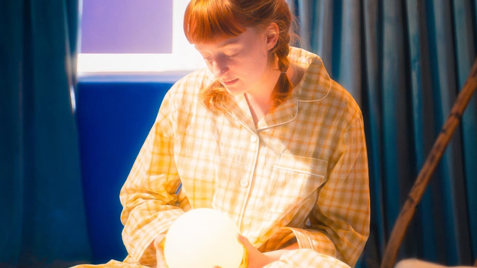 Die Künstlerin Emma Harmer im Schlafanzug sitzt auf einem Bett und hält eine leuchtende Kugel in einem abgedunkelten Raum, im Hintergrund ein dunkelblauer Vorhang und ein Stock.