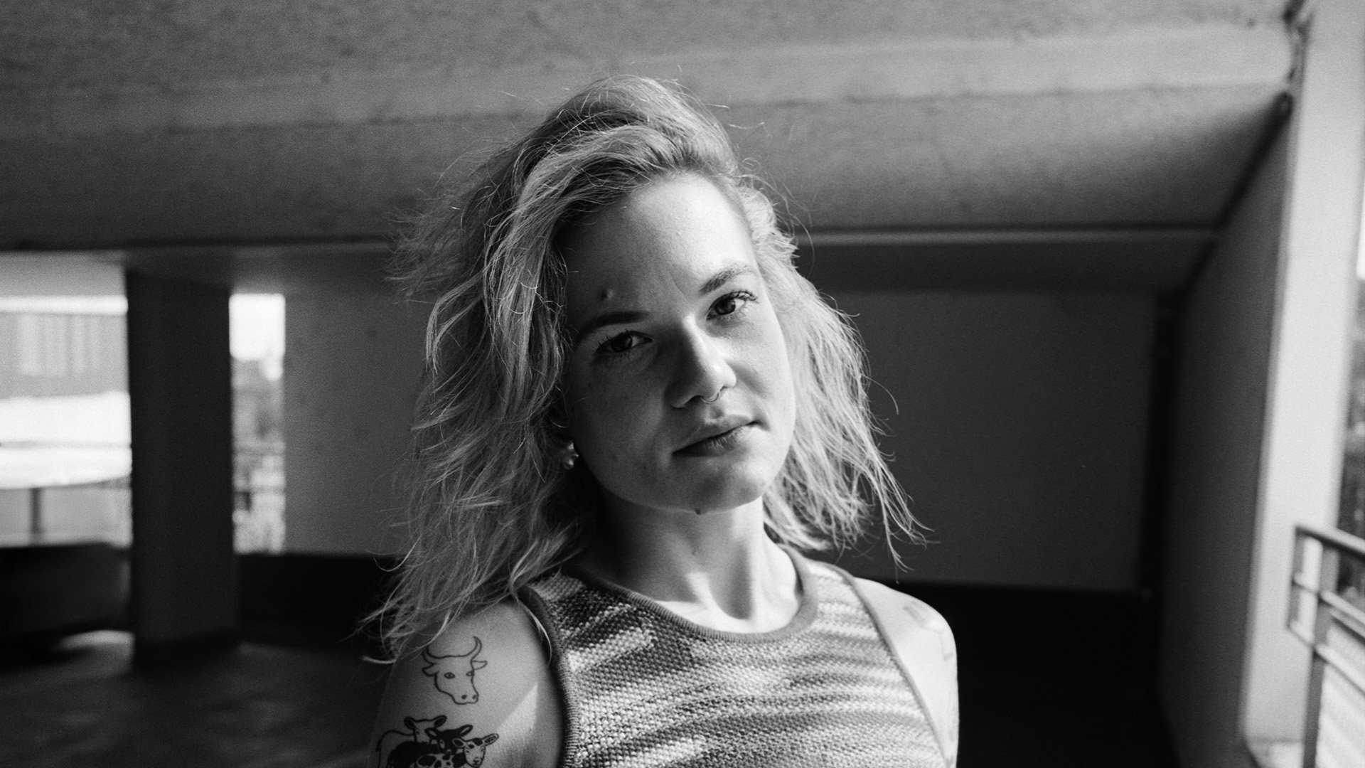 Schwarz-Weiß-Porträt von Avec mit schulterlangem Haar und sichtbaren Tattoos am Oberarm in einer urbanen Innenraumkulisse.