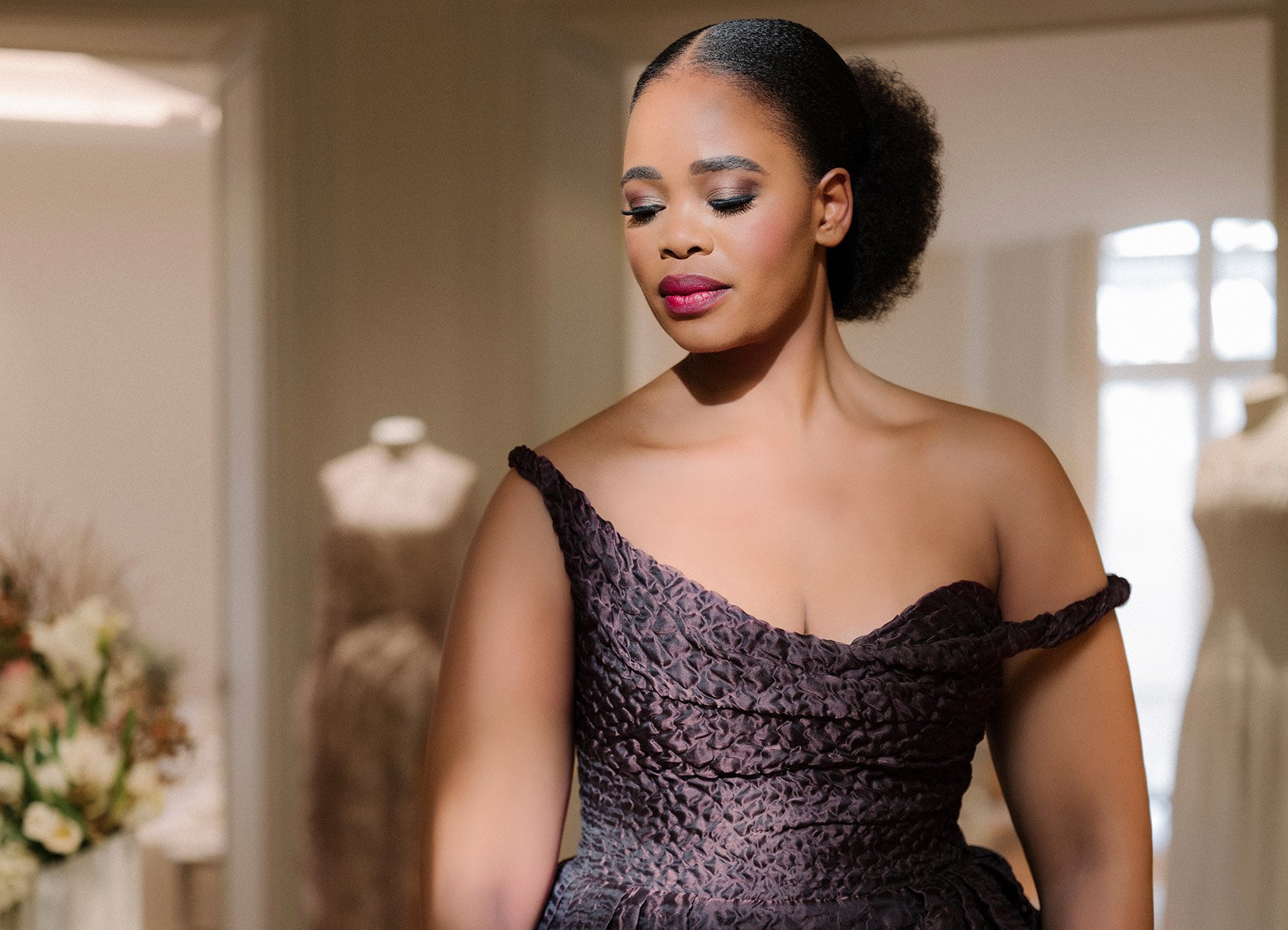 Porträt der Opernsängerin Pretty Yende in einem eleganten braunen Kleid – im Vordergrund, mit verschwommenem Raum im Hintergrund